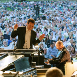 Photo of a past Pasadena POPS Summer concert, with conductor leading orchestra. A sea of delighted audience members is in the background.