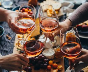 Four hands holding glasses of wine toast over a spread of cheese and fruits.