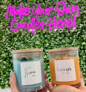 Two palms holding custom made candles with a neon sign in the background that states: Make Your Own Candles Here