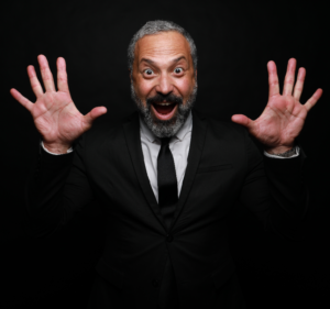 Photo of comedian Ahmed Ahmed wearing a black tie and suit in front of a black background.