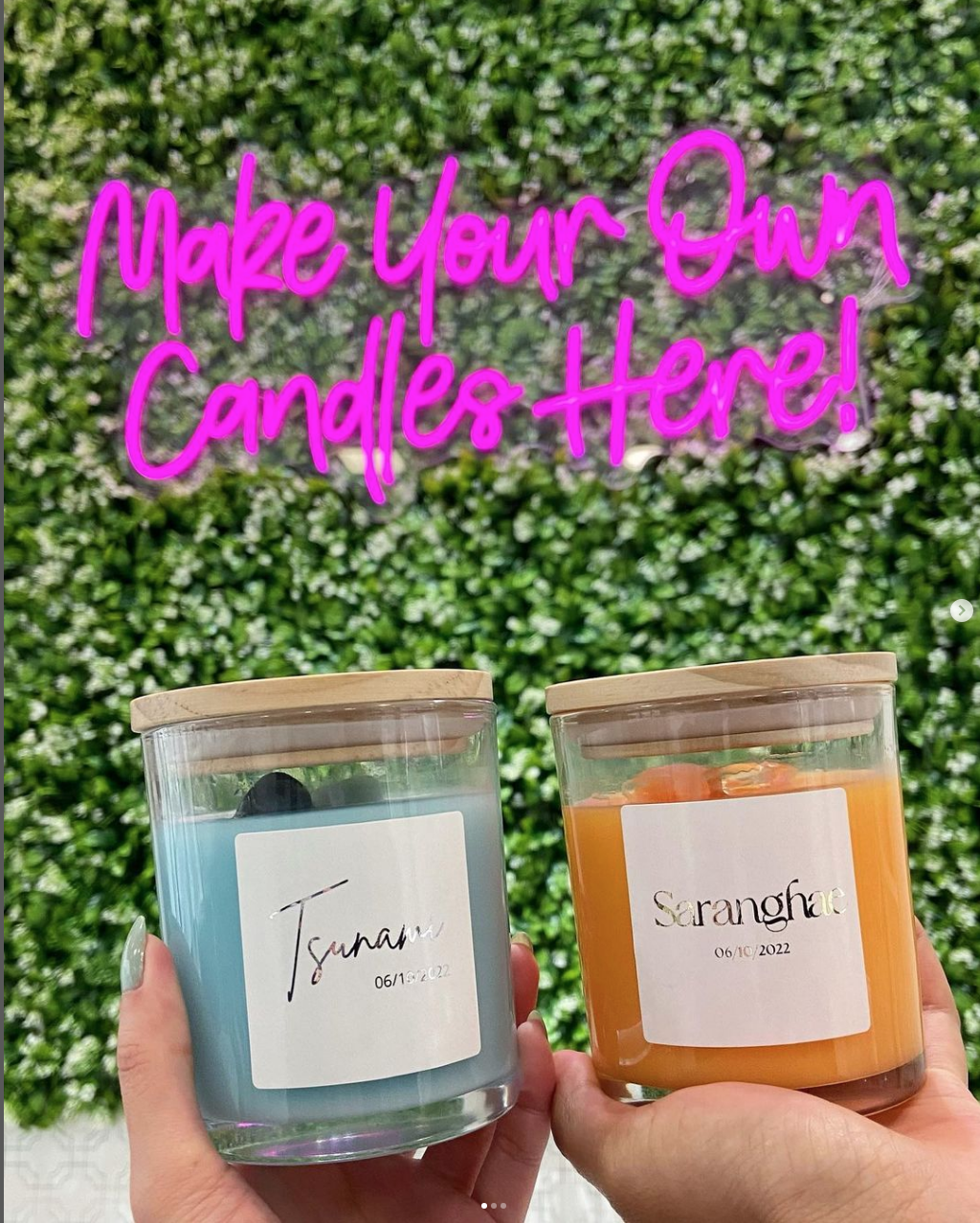 Two custom candles held below a pink neon sign that states: Make Your Own Candles Here