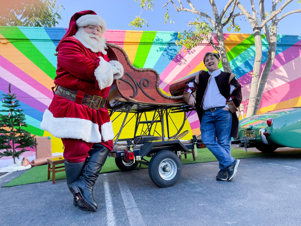 Santa stands next to his sleigh in front of the rainbow mural at Southern California Children's Museum