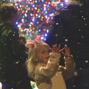 Young girl holds her hands out to touch the flurry of faux snow falling at the tree lighting at Playhouse Village