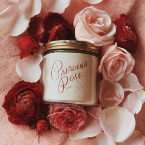 A Little Swiss Chalet candle labeled Pasadena Rose sits on a bed of pink and red roses