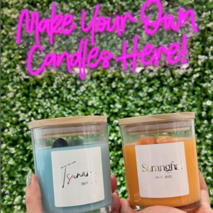 Two hands hold up two custom candles in front of a greenery backdrop with a neon sign that says Make Your Own Candles Here! This is at Banter & Bliss