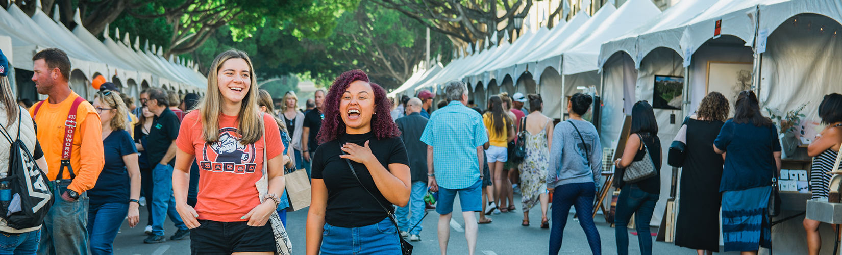 Two young women laugh as they stroll the art fair on a tree lined street, with artist booths and festival attendees in the background.