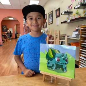 Young boy proudly stands next to his painting of Pokemon on an art easel.