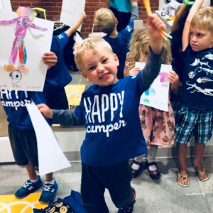 Young child wearing a Happy Camper shirt smiles as he holds up a craft he created at camp.