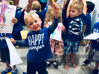 Young boy wearing a Happy Camper shirt smiles as he holds up an art & craft he created at camp