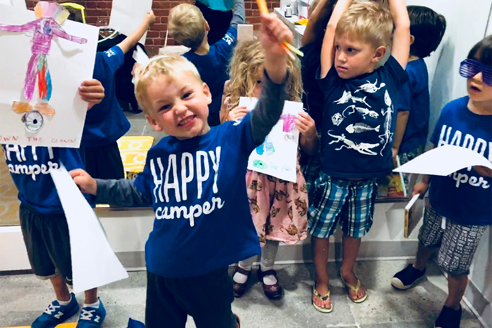 Young boy wearing a Happy Camper shirt smiles as he holds up an art & craft he created at camp