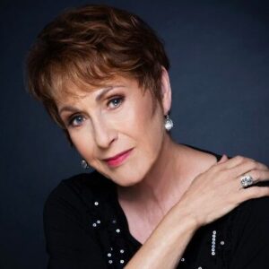 Photograph of Amanda McBroom wearing a black top with sparkling details, on a dark blue background.